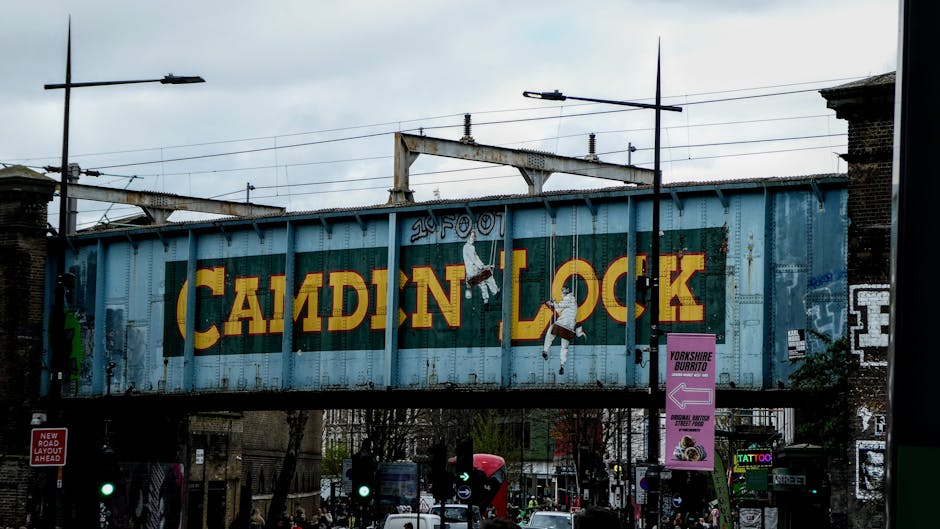 A large, weathered railway bridge painted in light blue spans across a busy street, prominently featuring the word 'Camden Lock' in bold, vintage-style yellow and red lettering. The bridge's metal sur