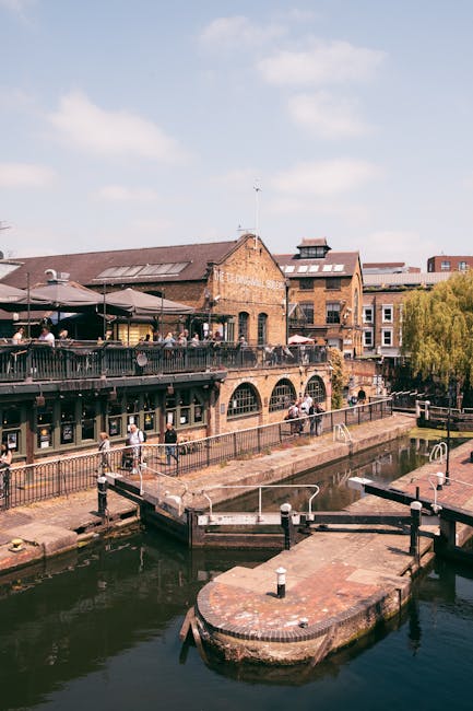 A large, weathered railway bridge painted in light blue spans across a busy street, prominently featuring the word 'Camden Lock' in bold, vintage-style yellow and red lettering. The bridge's metal sur
