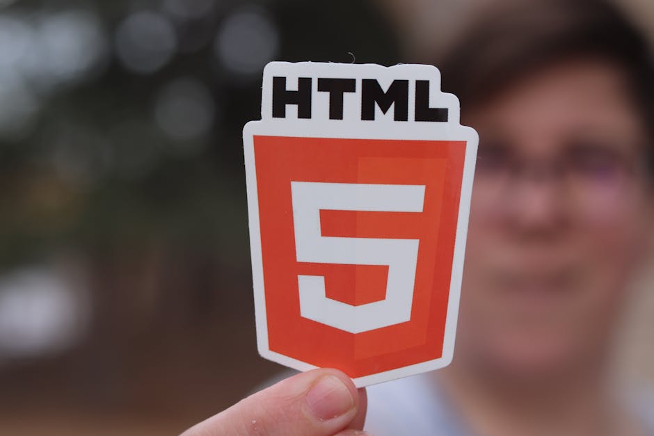 A person holding a cutout sticker with the HTML5 logo, featuring a large orange shield symbol with a white '5' in the center and the text 'HTML' above it. The individual is positioned outdoors, with a